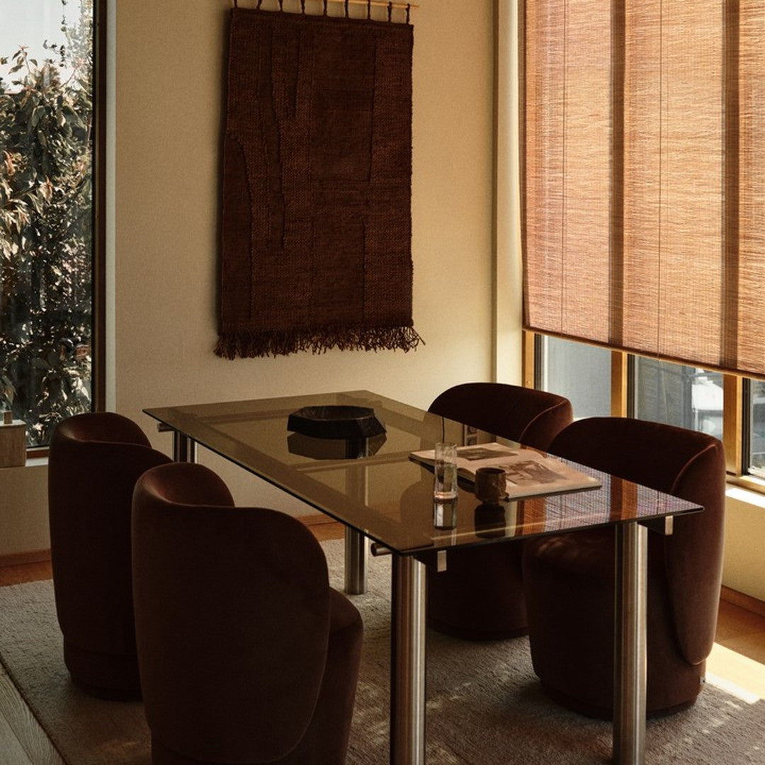 "Brown patterned wall hanging with warm wood tones, glass table, and rounded burgundy chairs in a cozy dining setting."