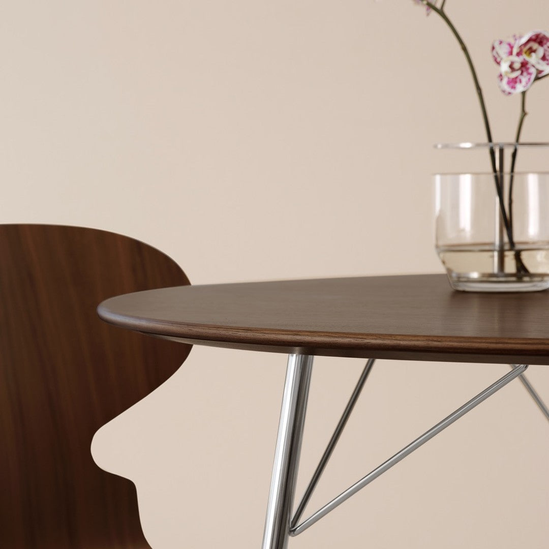 Brown wood table with a sleek round top, paired with a sculptural chair silhouette against a soft beige backdrop.