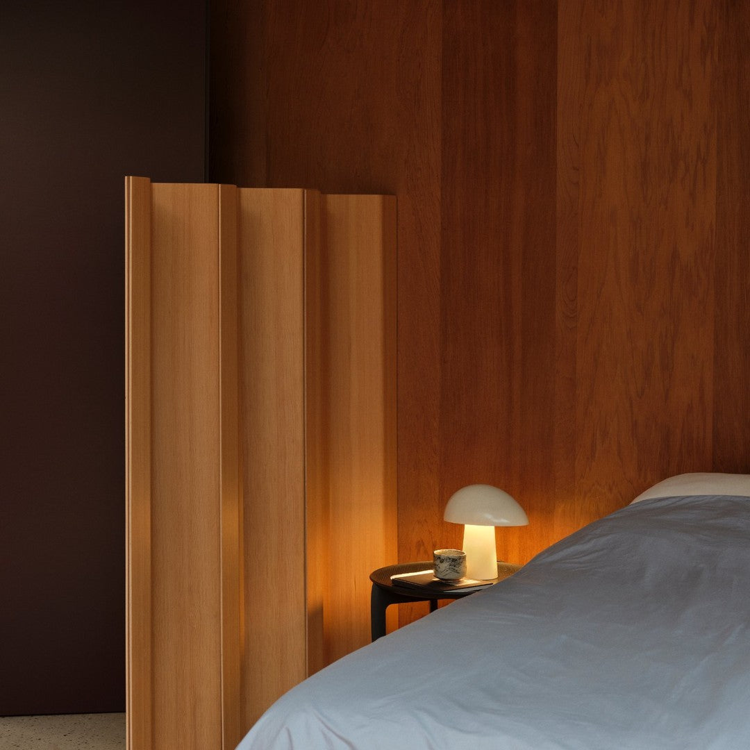 Wooden room divider in warm honey tones, blue bed linens, and a soft white lamp against a rich brown wall.