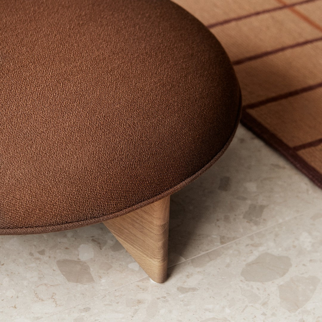Brown upholstered ottoman with wooden legs, set on a neutral terrazzo floor beside a grid-patterned rug.