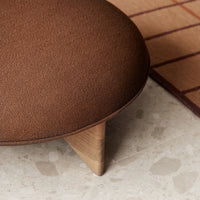 Brown upholstered ottoman with wooden legs, set on a neutral terrazzo floor beside a grid-patterned rug.