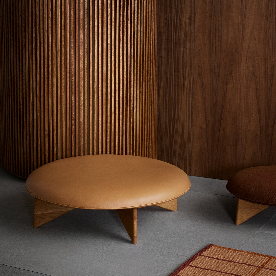 Modern round coffee table in warm tan leather with wooden legs, set against dark wood and neutral tones.
