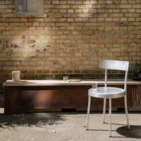 Silver metal chair on a rustic wooden bench, against a warm, textured brick wall. Natural light creates soft shadows.