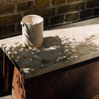White ceramic pitcher on a textured beige stone surface, with shadows and a rustic brown border.