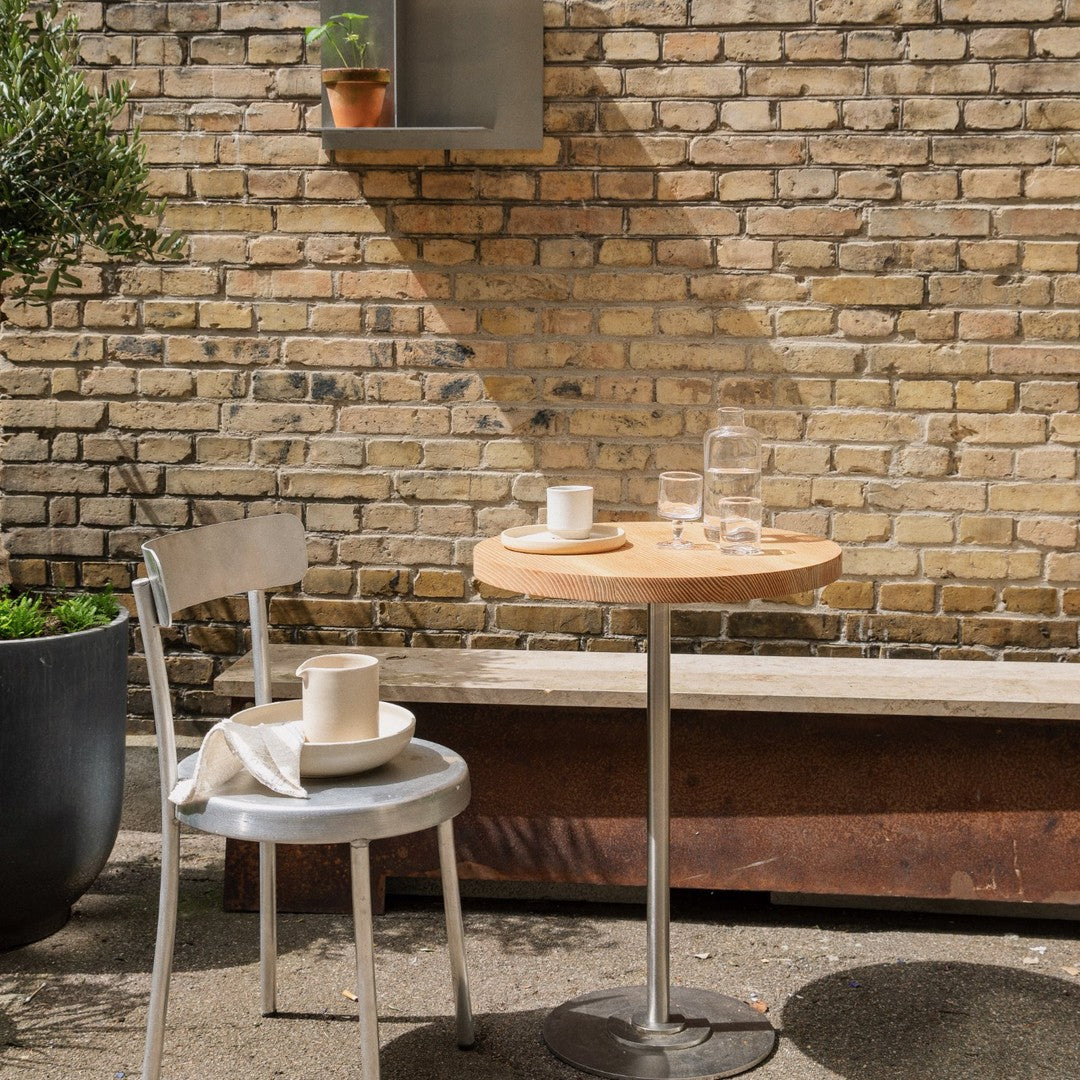 Light wood table and gray metal chair contrasted against a textured brick wall, featuring soft shadows and greenery.
