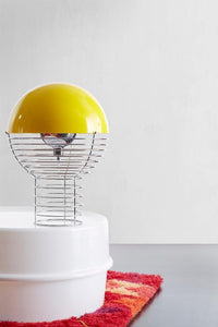 Yellow dome lamp with a wire base, set against a vibrant red and orange patterned rug on a white surface.