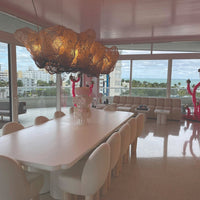 Modern dining room with a white table, soft beige chairs, and a unique bronze chandelier against pastel pink walls.