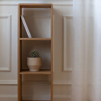 Natural wood bookshelf with a minimalist design, featuring a light beige ceramic pot and a subtle patterned curtain.