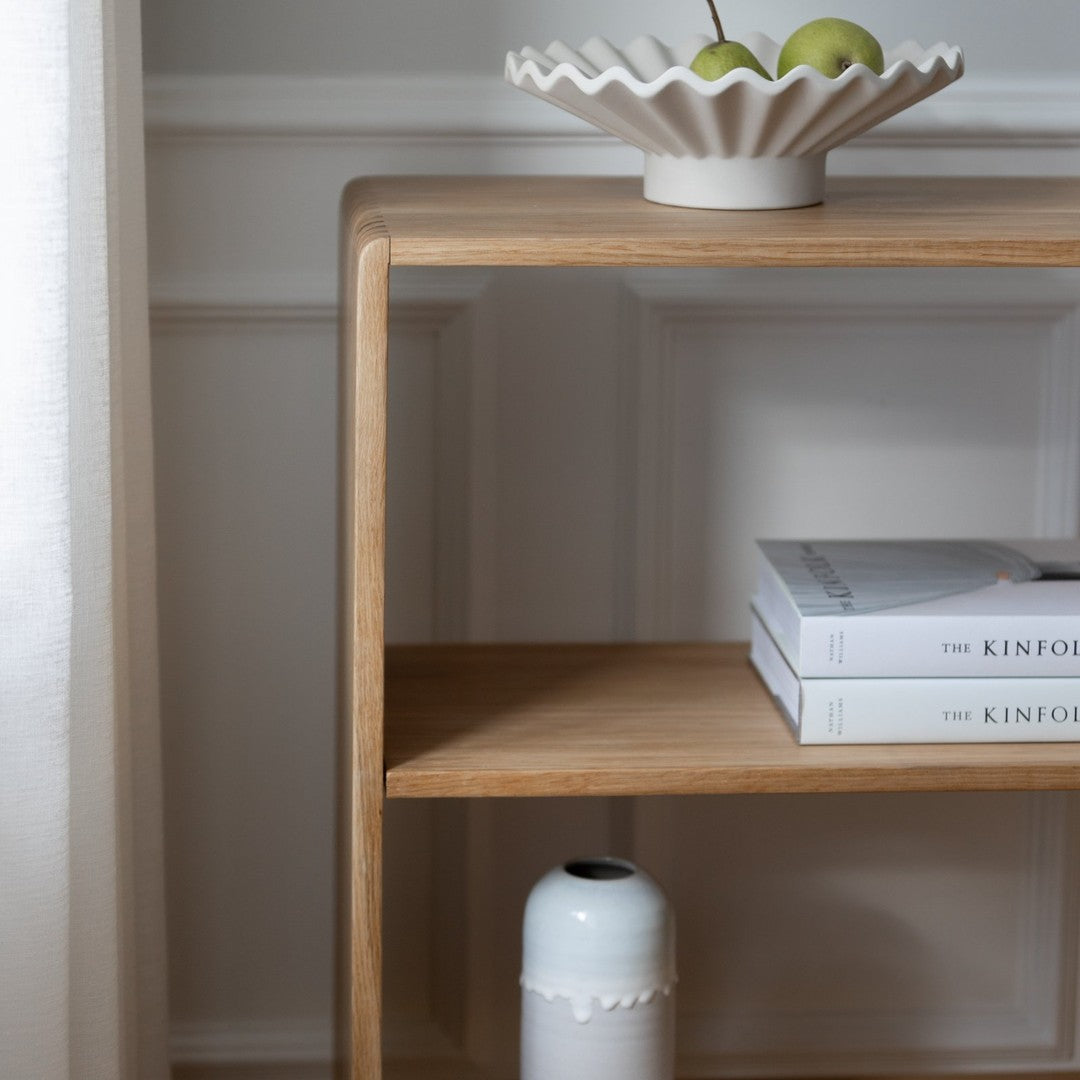 Light wood shelf featuring a white ribbed bowl, green apples, and stacked white books. Elegant, minimalist design.