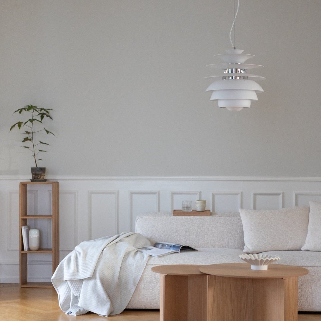 Modern white pendant light with layered design, set against a soft gray wall and natural wood furnishings.