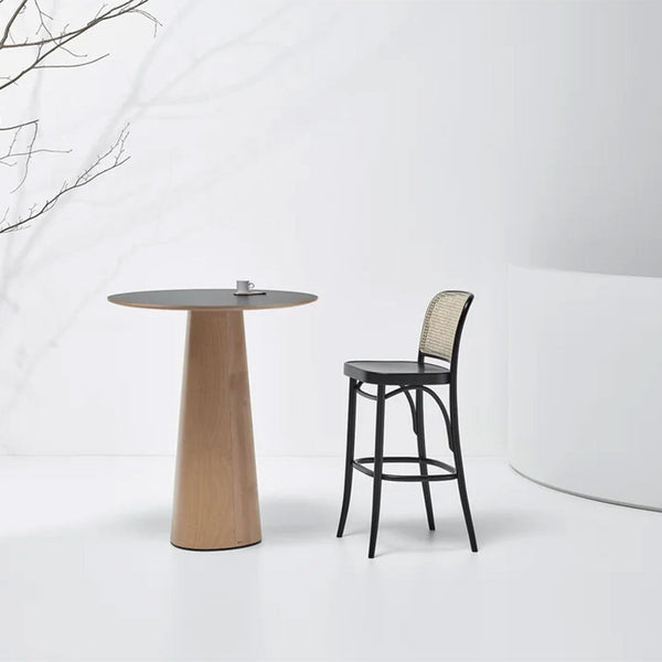 Light wood table with a black top beside a sleek black and rattan bar stool, set against a minimalist white backdrop.