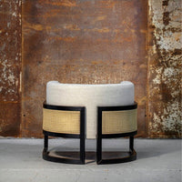 Round upholstered chair in soft cream fabric with a woven cane pattern on black frame, set against a rustic background.
