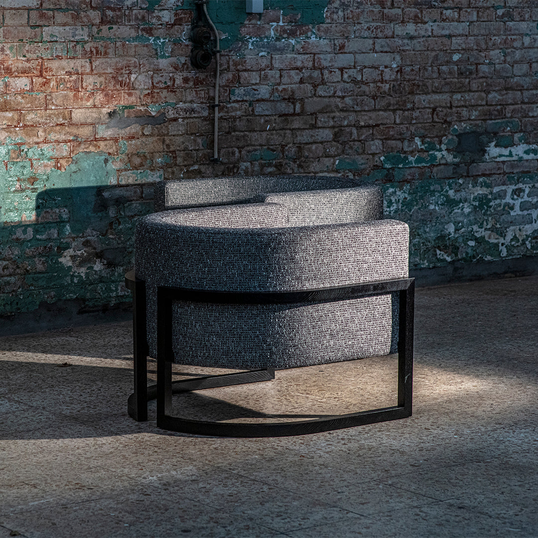 Gray upholstered chair with a curved shape and black metal frame, set against a textured brick background.