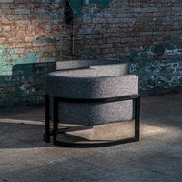 Gray upholstered chair with a curved shape and black metal frame, set against a textured brick background.