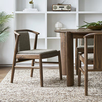 Wooden chair with textured gray upholstery on a neutral woven rug, set against a white shelving unit.