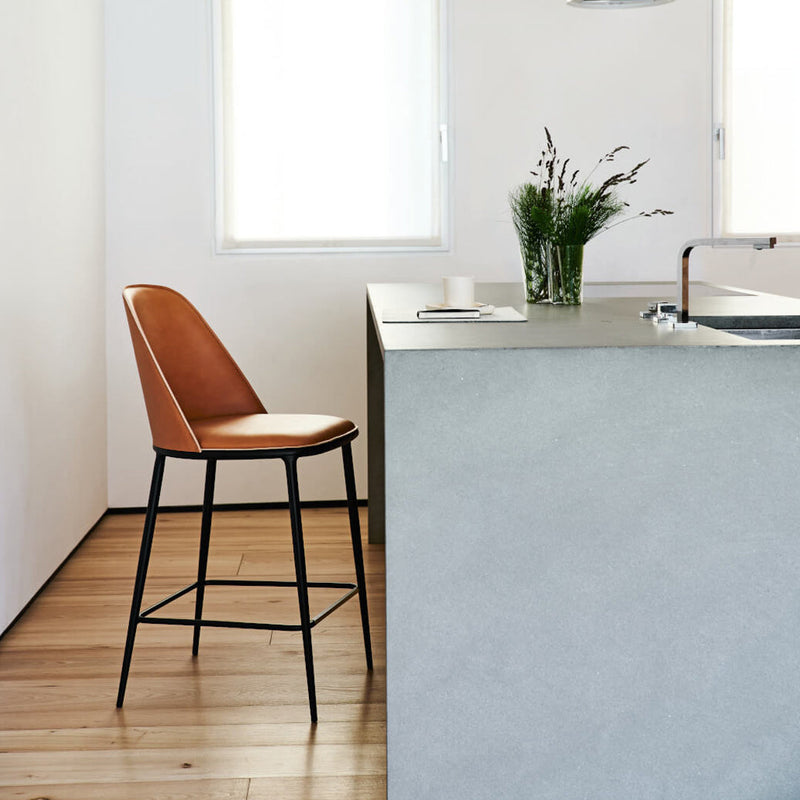 Tan leather bar stool with a sleek black metal frame, set against a light wooden floor and minimalist gray counter.