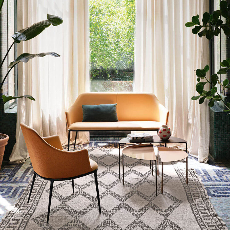 Orange sofa and armchair on a patterned rug with geometric designs, surrounded by green plants and light curtains.