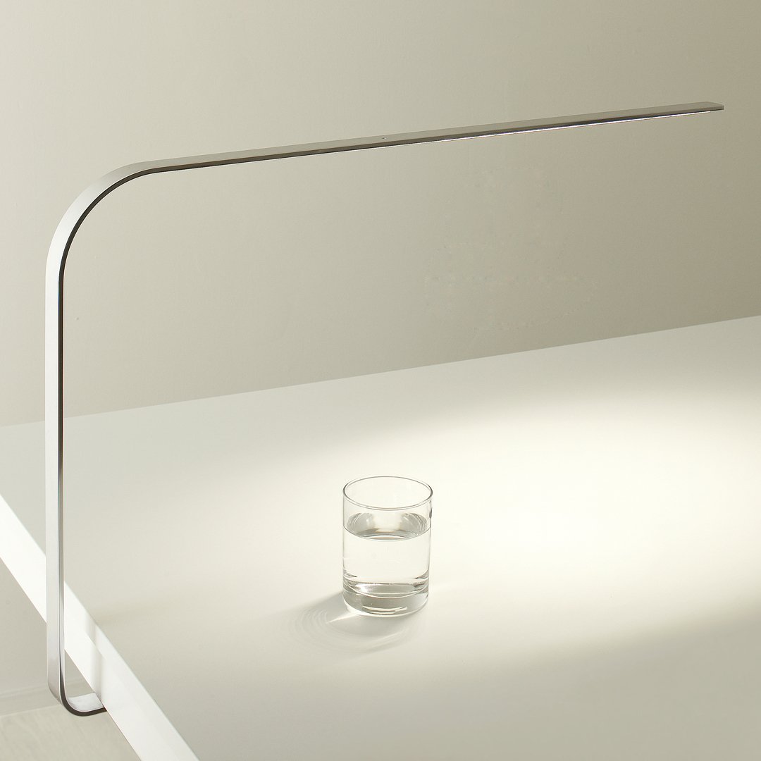 Sleek silver desk lamp with minimalist design casting warm light on a clear glass of water.