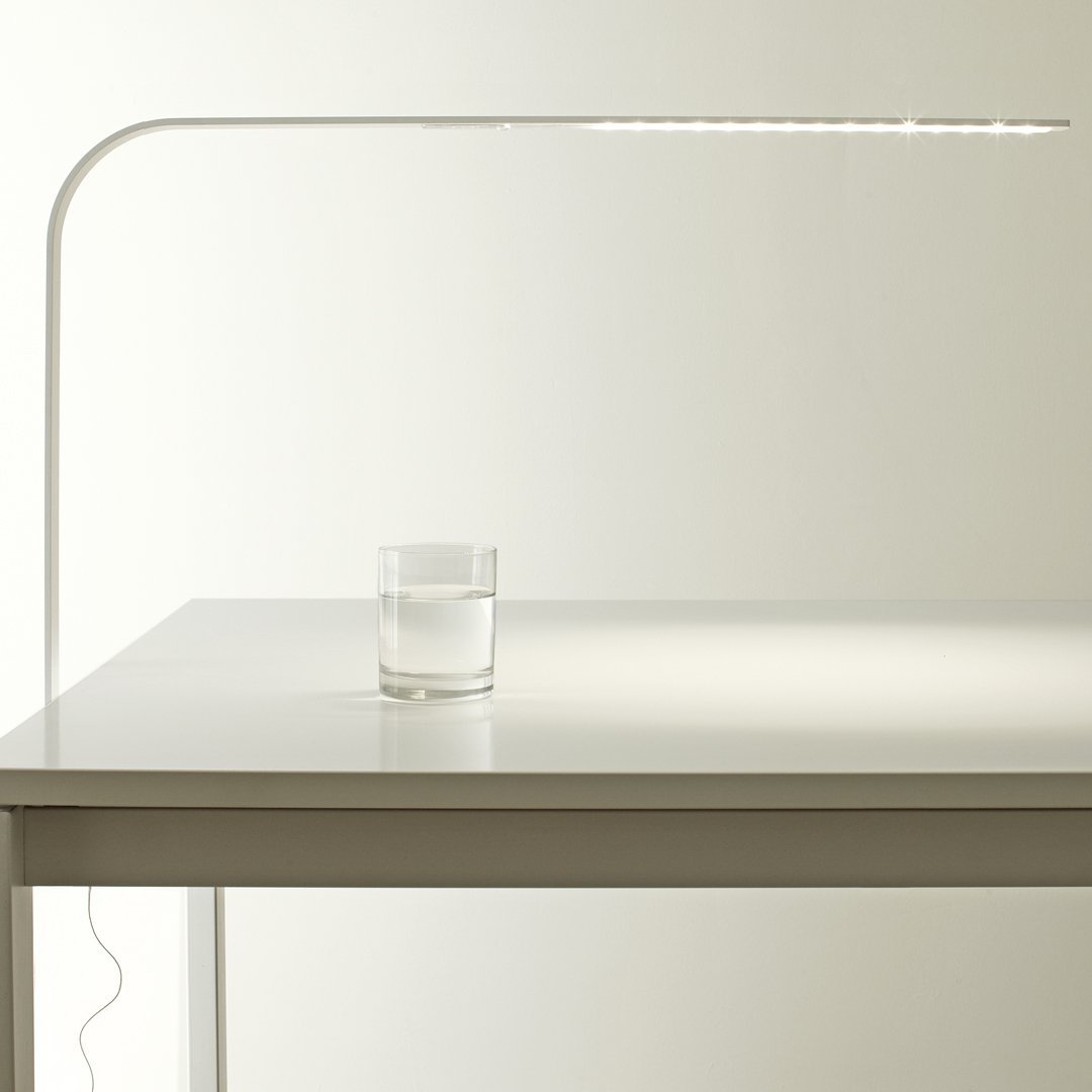 Sleek white desk lamp with a minimalist design, featuring a modern curved arm and a clear glass of water on a white table.