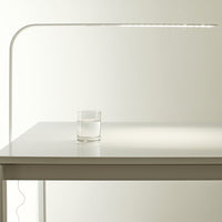 Sleek white desk lamp with a minimalist design, featuring a modern curved arm and a clear glass of water on a white table.
