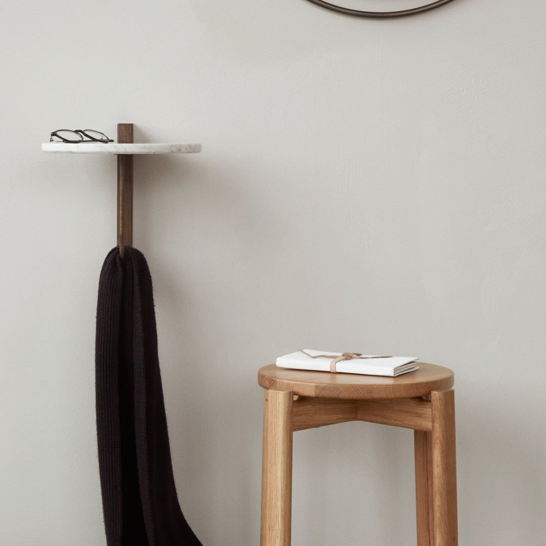 Minimalist wooden stool with a natural finish and black fabric draped on a simple white and brown shelf against a neutral wall.