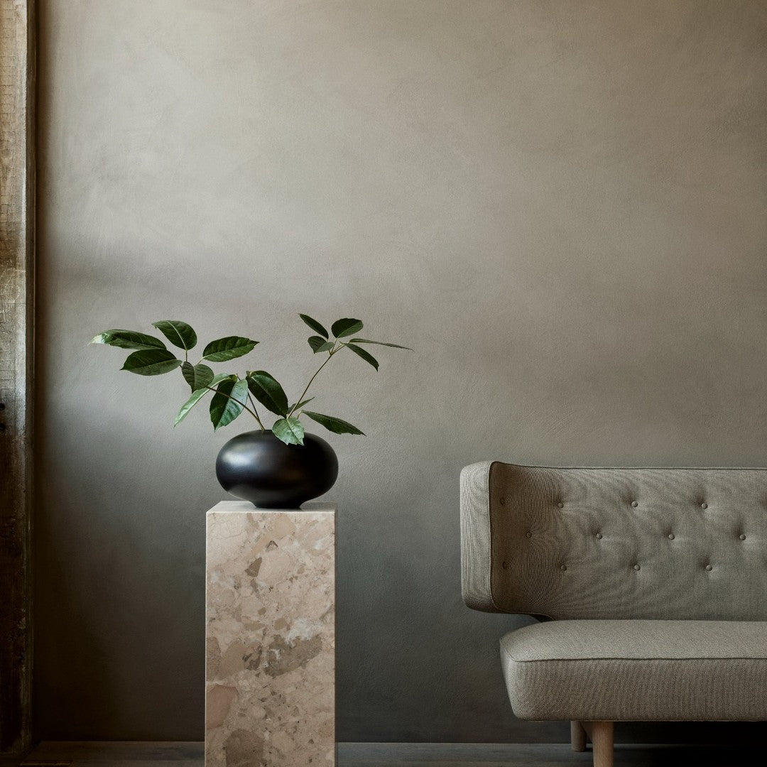 Elegant green leaves in a black vase atop a beige marble pedestal, complemented by a light gray tufted sofa against a muted backdrop.