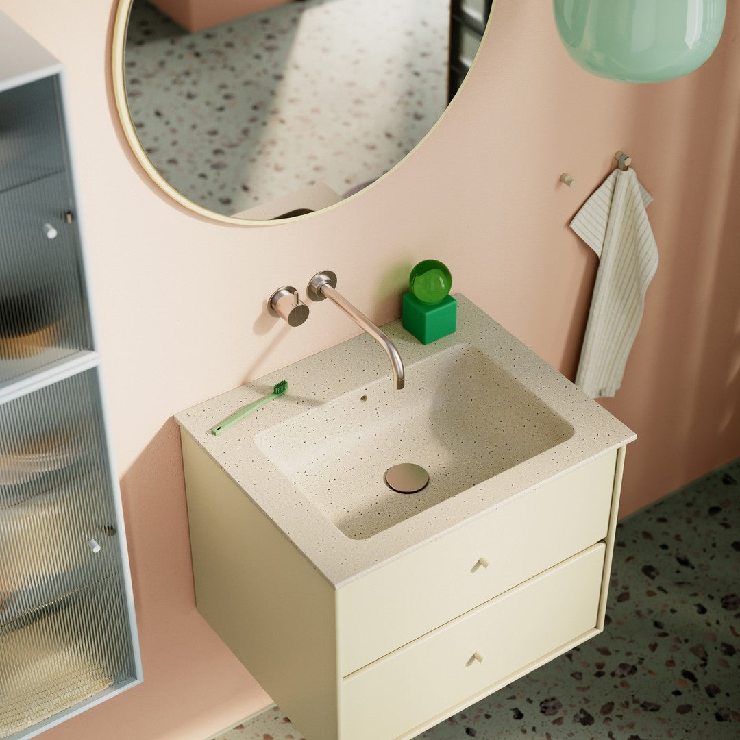 "Light yellow bathroom vanity with speckled countertop, pink walls, green accents, and a reflective round mirror."