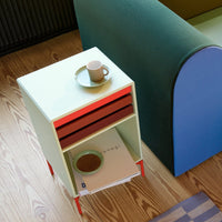 Mint green nightstand with red drawers and orange legs, complemented by a cozy blue sofa and patterned floor.