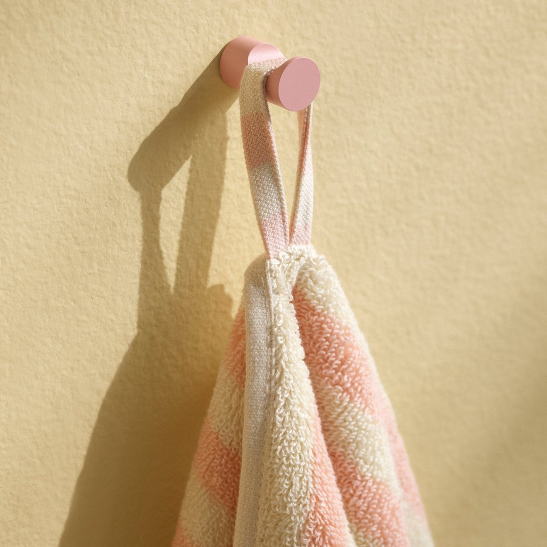 Soft beige towel with coral pink stripes, hanging from a pastel pink hook against a yellow wall.