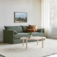 Green fabric sofa with rust-colored pillows, set on a beige rug, against a soft-lit neutral backdrop.