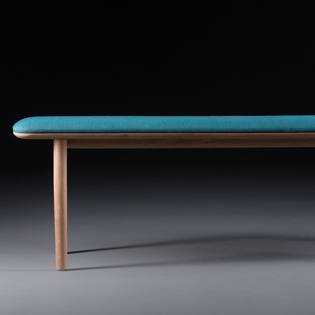 Teal upholstered bench with a smooth surface and wooden legs, set against a dark background.