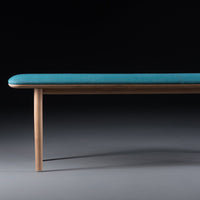 Teal upholstered bench with a smooth surface and wooden legs, set against a dark background.