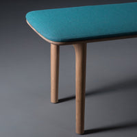 Teal upholstered bench with sleek wooden legs on a dark gray background. Modern minimalist design.