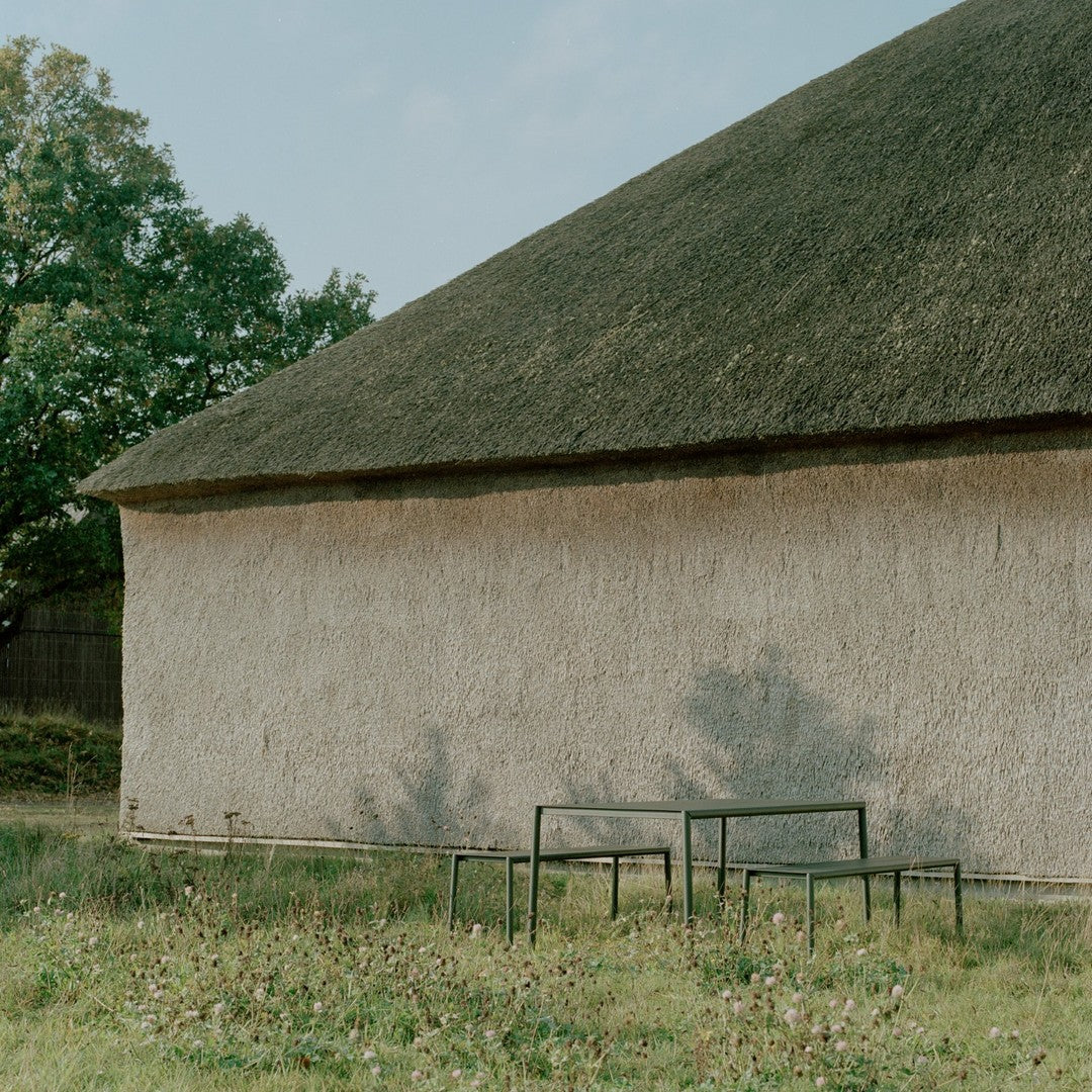 "Neutral-toned wall with a thatched roof and minimalist black metal tables against a grassy background."