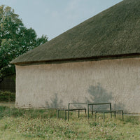 "Neutral-toned wall with a thatched roof and minimalist black metal tables against a grassy background."