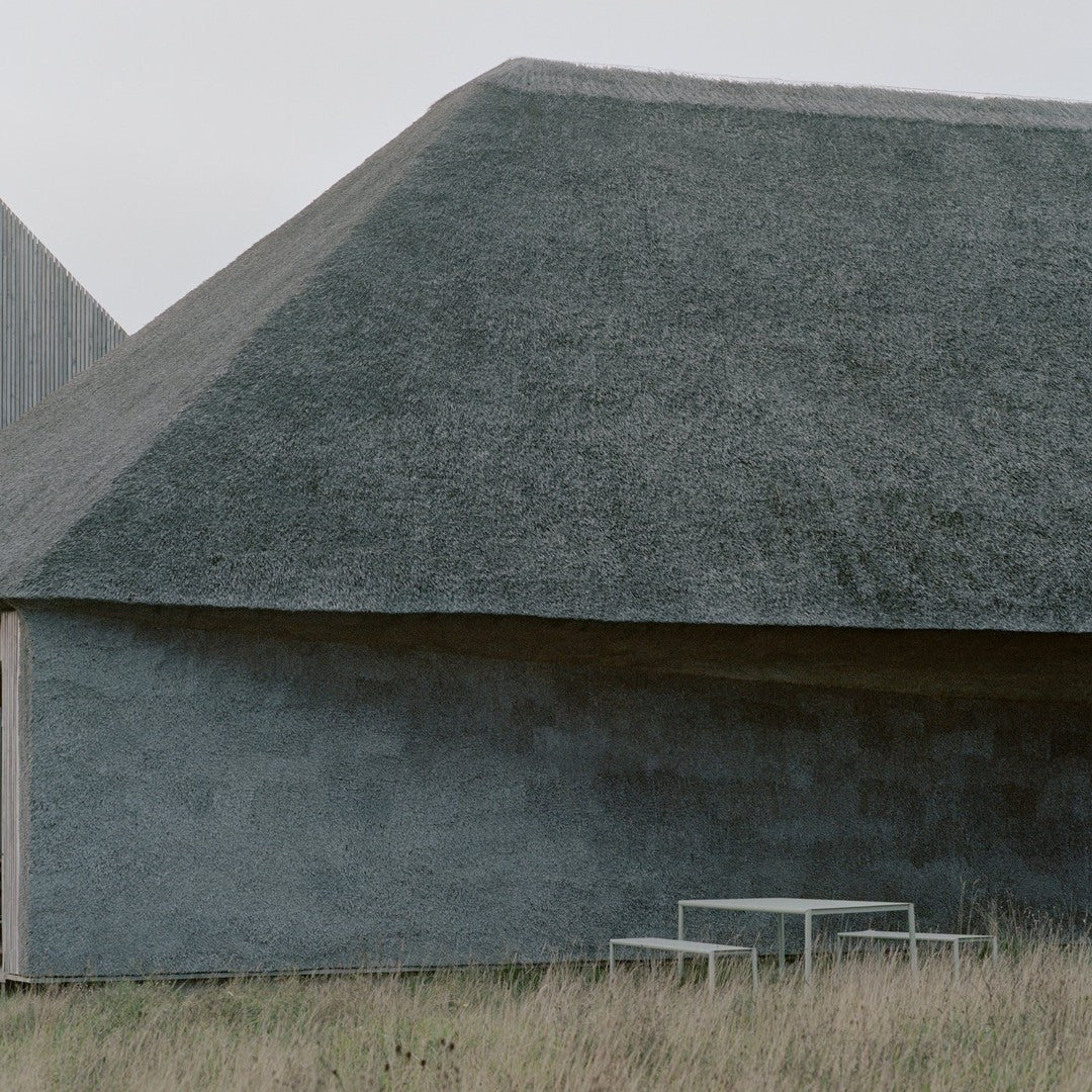 Gray textured building with a sloped roof, surrounded by green grass and a minimalist white table in the foreground.