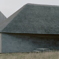 Gray textured building with a sloped roof, surrounded by green grass and a minimalist white table in the foreground.