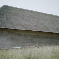 Grey thatched roof on a minimalist building with a textured stone wall, surrounded by green grass.