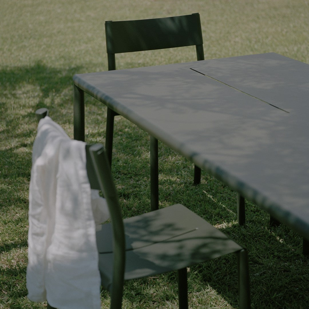 Green outdoor dining table and chairs with a white towel draped over a chair, set on a grassy background.