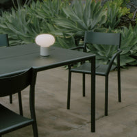 Green outdoor dining set with minimalist design and a round white lamp, set against lush greenery.