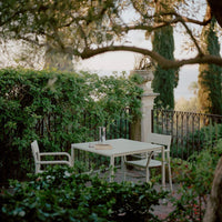 White outdoor table and chairs surrounded by lush green foliage and trees, creating a serene garden ambiance.
