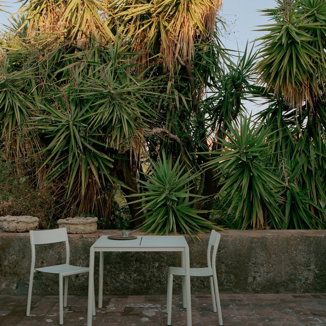 "Minimalist white outdoor table and chairs surrounded by lush green tropical foliage."