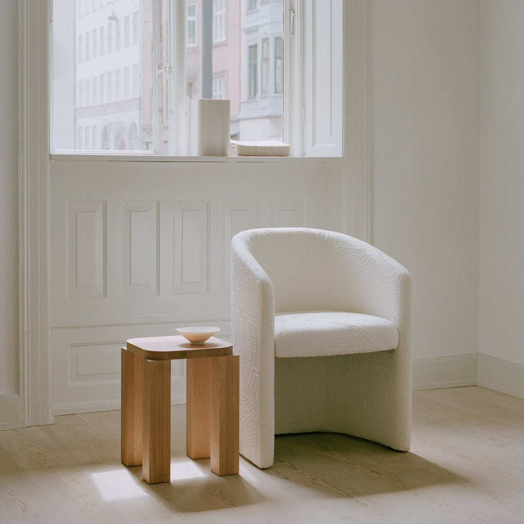 Elegant white upholstered chair with embossed pattern beside a natural wood side table in a bright room setting.