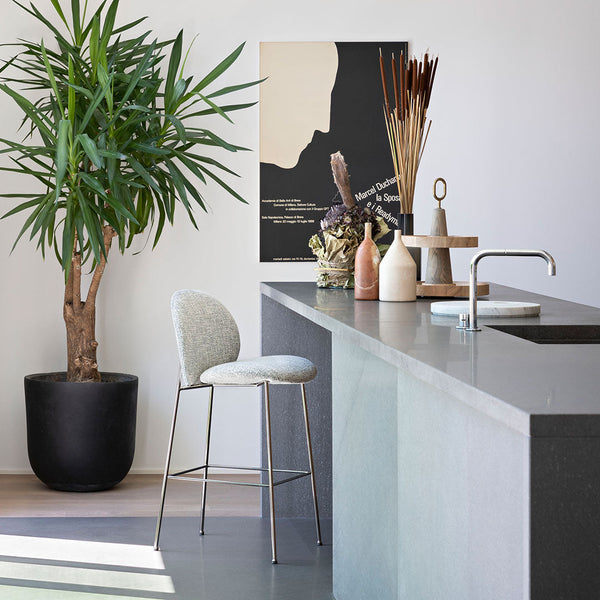 Light gray bar stool with subtle texture, set against neutral tones with natural green foliage and minimalist decor.