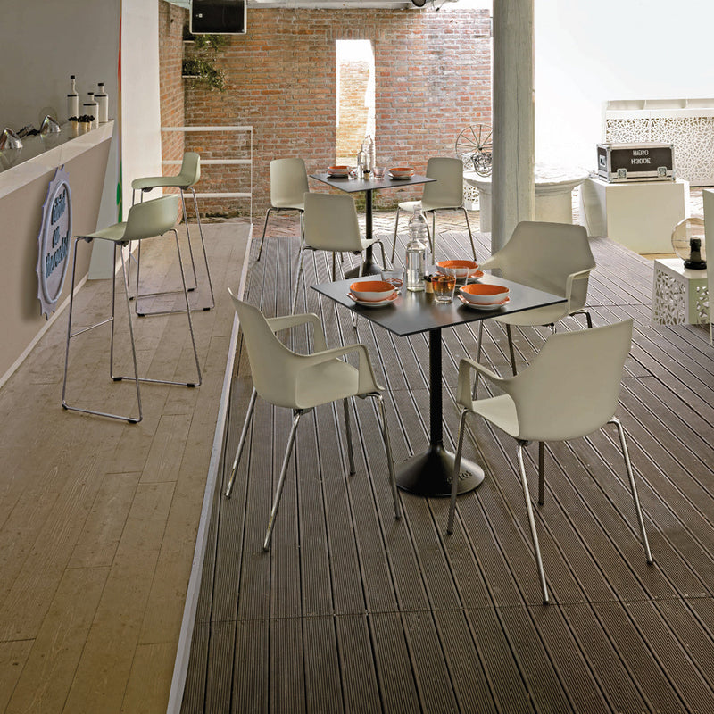 Modern outdoor space featuring white chairs and black tables against a rustic brick backdrop and wooden flooring.