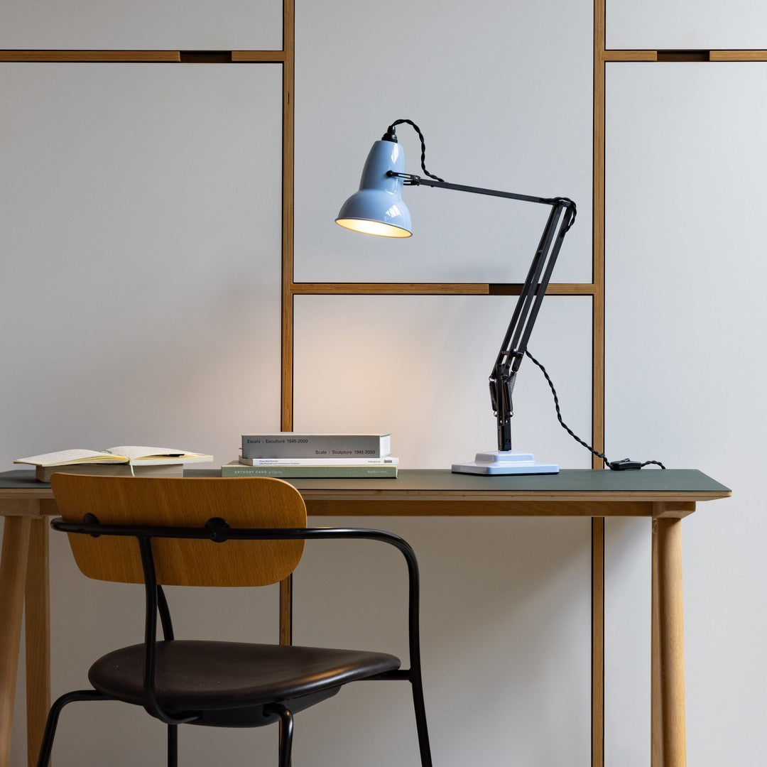 Blue desk lamp with a black arm on a green tabletop, surrounded by neutral-toned furniture and light gray walls.