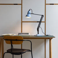 Blue desk lamp with a black arm on a green tabletop, surrounded by neutral-toned furniture and light gray walls.