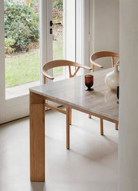 Light gray table with natural wood legs, surrounded by minimalist wooden chairs in a sunny, green garden setting.