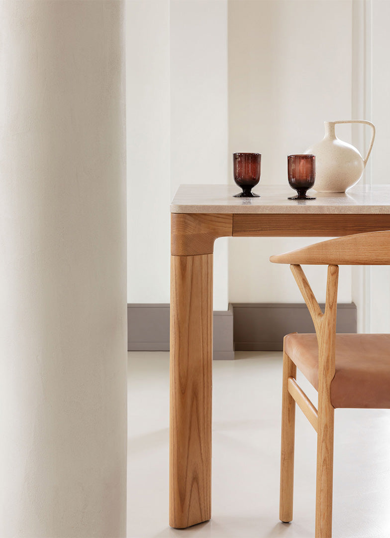 Light wood table with a natural finish, soft beige chair, and dark glassware, set against neutral-toned walls.