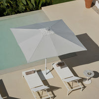 White square patio umbrella paired with minimalist white lounge chairs beside a tranquil pool. Bright and airy design.